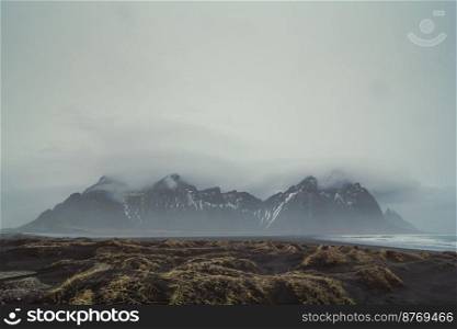 Black beach with dry grass landscape photo. Beautiful nature scenery photography with mountains on background. Idyllic scene. High quality picture for wallpaper, travel blog, magazine, article. Black beach with dry grass landscape photo