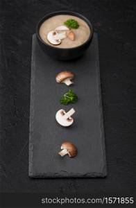Black bowl plate of creamy chestnut champignon mushroom soup on black background with black stone board and fresh mushrooms.