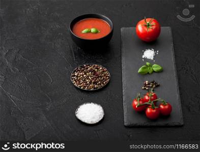 Black bowl plate of creamy tomato soup on black background with stone chopping board and raw tomatoes, pepper and salt. Space for text