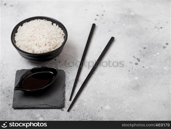 Black bowl with boiled organic basmati jasmine rice with black chopsticks and sweet soy sauce on stone mat on light kitchen table background.