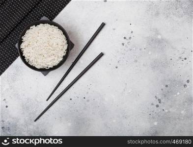 Black bowl with boiled organic basmati jasmine rice with black chopsticks and sweet soy sauce on stone mat on light kitchen table background.