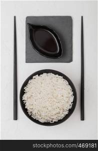 Black bowl with boiled organic basmati jasmine rice with black chopsticks and sweet soy sauce on stone mat on white.