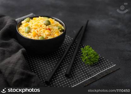 Black bowl with boiled organic basmati vegetable rice with black chopsticks on placemat with linen towel on black background.. Yellow corn and green peas with paprika slices.