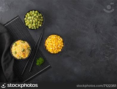 Black bowl with boiled organic basmati vegetable rice with black chopsticks on placemat with linen towel on black background.. Yellow corn and green peas with paprika slices.