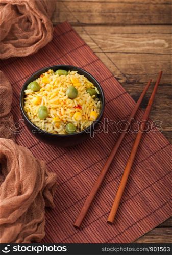 Black bowl with boiled organic basmati vegetable rice with wooden chopsticks on brown placemat with linen towel. Yellow corn and green peas with paprika slices.