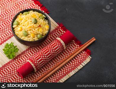 Black bowl with boiled organic basmati vegetable rice with wooden chopsticks on red bamboo placemat. Yellow corn and green peas with paprika.