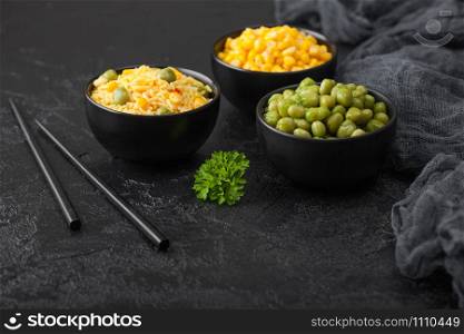 Black bowls with boiled organic basmati vegetable rice, yellow corn and peas with black chopsticks on black stone background.