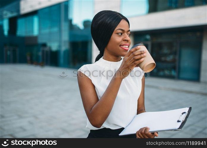 Black business girl grinks coffee from cardboard cup outdoors, office building on background. Black businesswoman in white blouse on working break. Business girl grinks coffee from cardboard cup