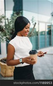 Black business woman with cardboard coffee cup uses mobile phone against office building on background. Smiling businesswoman in skirt and white blouse