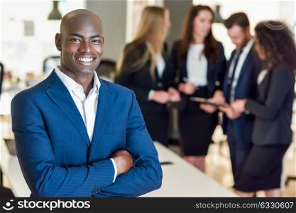Black businessman leader looking at camera in modern office with multi-ethnic businesspeople working at the background. Teamwork concept. African man wearing blue suit.