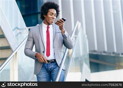 Black Businessman sending voice note with his smartphone near an office building. Man with afro hair.. Black Businessman using a smartphone near an office building