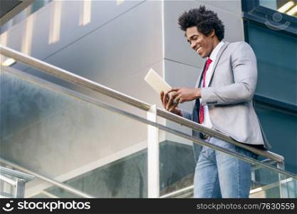Black Businessman using a digital tablet sitting near an office building. Man with afro hair.. Black Businessman using a digital tablet sitting near an office building.
