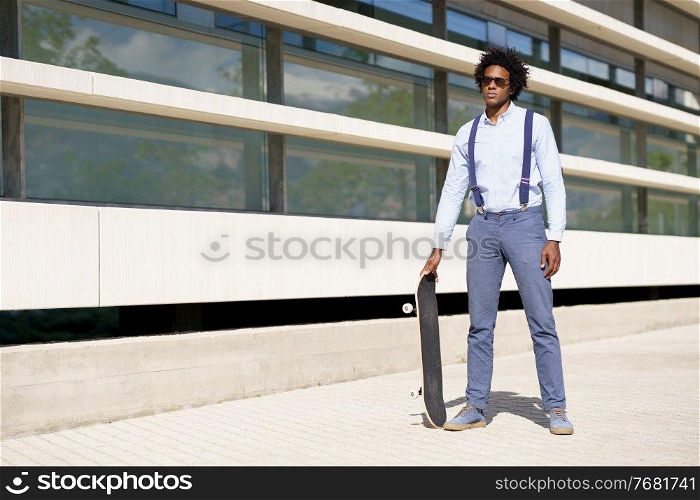 Black businessman with afro hairstyle standing next to an office building with a skateboard and sunglasses.. Black male worker standing next to an office building with a skateboard.
