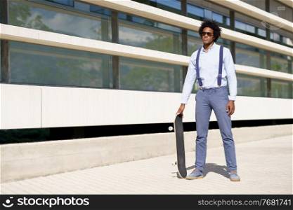 Black businessman with afro hairstyle standing next to an office building with a skateboard and sunglasses.. Black male worker standing next to an office building with a skateboard.