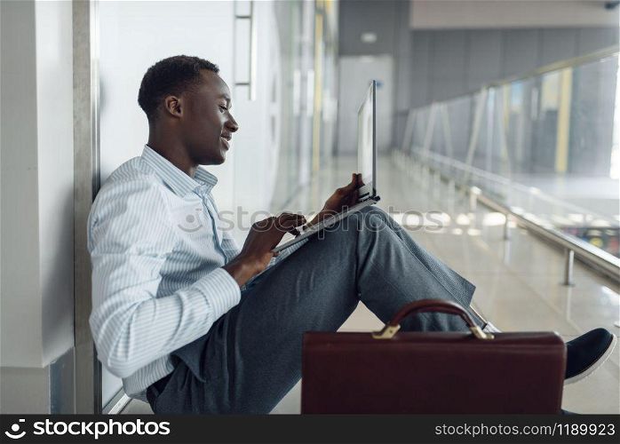 Black businessman with briefcase and laptop sitting on the floor in office hallway. Tired business person relax in corridor, black man in formal wear. Black businessman with laptop sitting on the floor