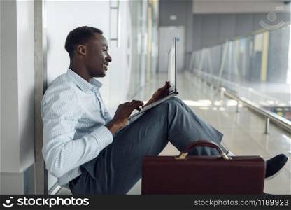 Black businessman with briefcase and laptop sitting on the floor in office hallway. Tired business person relax in corridor, black man in formal wear. Black businessman with laptop sitting on the floor