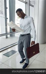 Black businessman with briefcase reading newspaper, mall. Successful business person, black man in formal wear, shopping center. Black businessman with briefcase reading newspaper