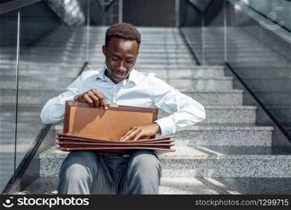 Black businessman with briefcase sitting on stairs, mall. Successful business person, black man in formal wear, shopping center. Black businessman with briefcase sitting on stairs