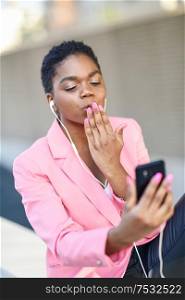 Black businesswoman sitting outdoors speaking via videoconference with her smartphone. African american female wearing suit with pink jacket.. Black businesswoman sitting outdoors speaking via videoconference with her smart phone.