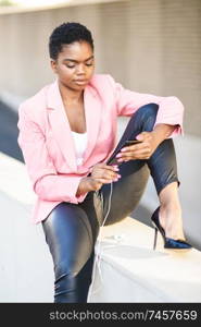 Black businesswoman sitting outdoors using smartphone with earphones. African american female wearing suit with pink jacket.. Black businesswoman sitting outdoors using smartphone with earphones