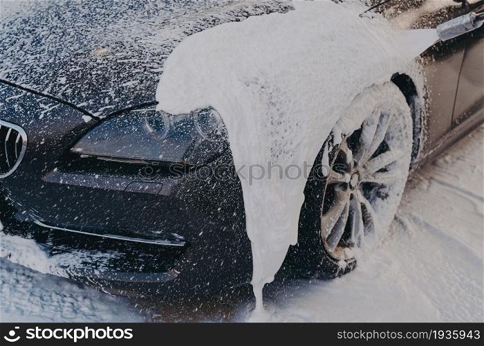Black car in white soap foam at washing station, professional vehicle cleaning with chemical detergent. Carwashing and detailing concept. Black car in white soap foam at washing station