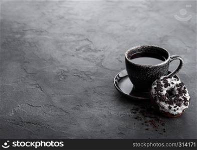 Black coffee cup with saucer and doughnut with black cookies on black stone kitchen table background. Space for text.