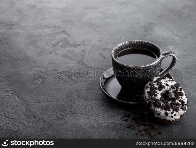 Black coffee cup with saucer and doughnut with black cookies on black stone kitchen table background. Space for text.
