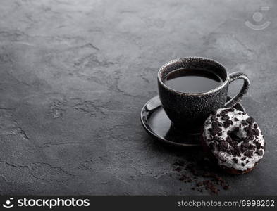 Black coffee cup with saucer and doughnut with black cookies on black stone kitchen table background. Space for text.