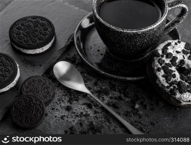 Black coffee cup with saucer and doughnut with black sandwich cookies on black stone kitchen table background. Space for text. Breakfast snack.