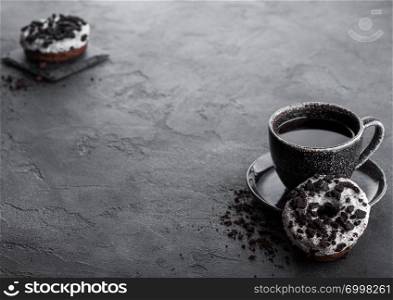 Black coffee cup with saucer and doughnuts with black cookies on black stone kitchen table background. Space for text.