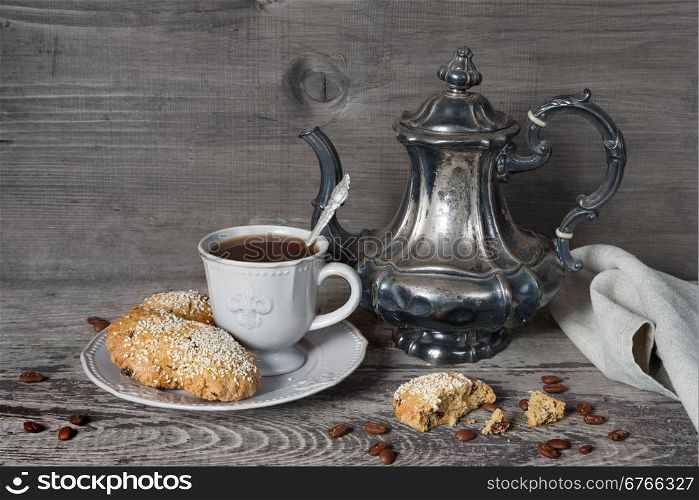 Black coffee in a stylized Victorian cup and oatmeal cookies sprinkled with sesame seeds on a porcelain plate as well as old silver coffee pot and gray linen napkin on a background of old unpainted boards