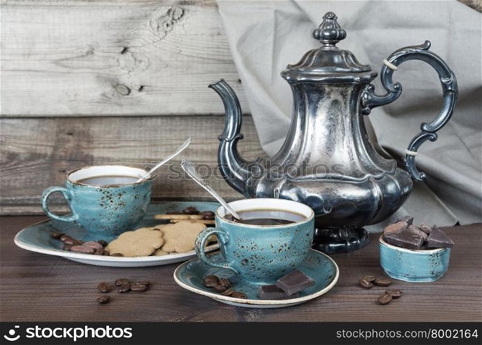 Black coffee in blue vintage cup, cookies, chocolate and antique silver coffee pot on a old dark wooden boards