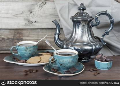 Black coffee in blue vintage cup, cookies, chocolate and antique silver coffee pot on a old dark wooden boards
