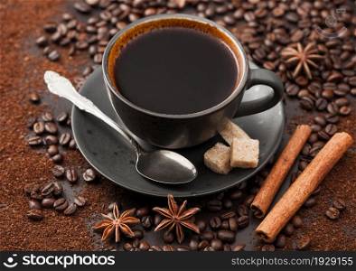 Black coffee in ceramic cup with cinnamon and cane sugar with anise star and silver spoon on ground coffee and beans background.