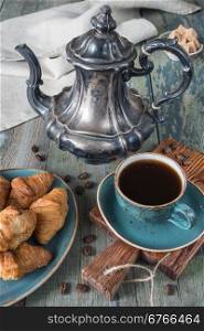 Black coffee in vintage cup, croissants on an old old blue plate and antique silver coffee pot on a old dark wooden boards