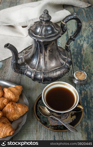 Black coffee in vintage cup, croissants on an old tin plate and antique silver coffee pot on a old dark wooden boards