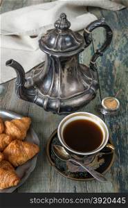 Black coffee in vintage cup, croissants on an old tin plate and antique silver coffee pot on a old dark wooden boards