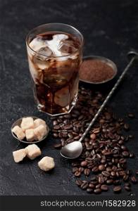 Black coffee with ice cubes and fresh milk with beans and cane sugar with ground coffee and long spoon on black background.