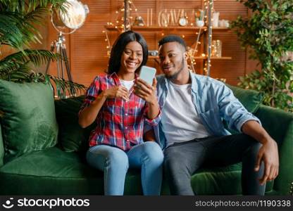 Black couple sitting on sofa and looking on mobile phone at home. Happy african love couple leisures in their house, cheerful family relaxing together