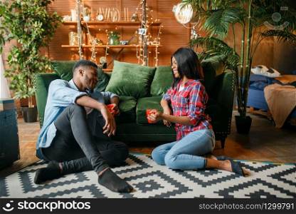 Black couple sitting on the floor and drinks coffee against a couch at home. Happy african love couple leisures in their house, cheerful family relaxing together in the morning. Black couple sitting on floor and drinks coffee