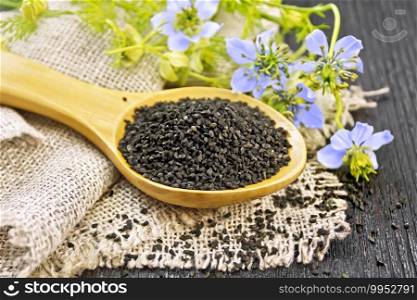 Black cumin seeds in a spoon on sacking, kalingi twigs with blue flowers and green leaves on a dark wooden board background