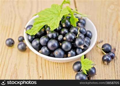 Black currants in a white bowl with green leaves on a wooden boards background