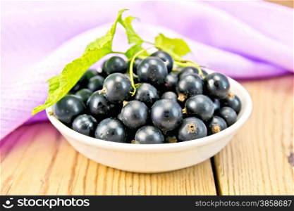 Black currants in a white bowl with green leaves, purple cloth on a wooden boards background