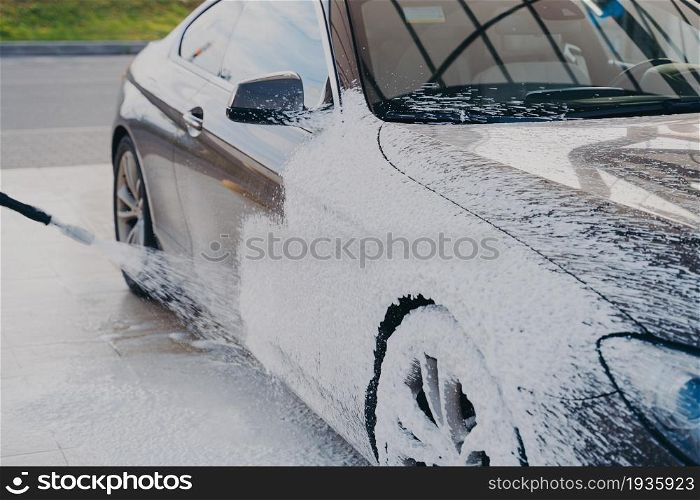 Black dirty car in white soap foam at car wash service station, vehicle cleaning with pressure washer and detergent. Washing process of auto exterior surface. Black dirty car in white soap foam at car wash service station