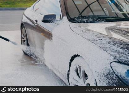 Black dirty car in white soap foam at car wash service station, vehicle cleaning with pressure washer and detergent. Washing process of auto exterior surface. Black dirty car in white soap foam at car wash service station