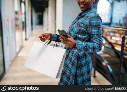 Black female person with phone and shopping bags in mall. Shopaholic in clothing store, consumerism lifestyle, fashion