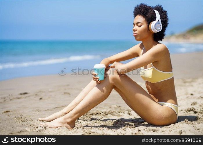 Black female sitting on the beach with wireless headphones, sipping a drink from a paper take-away cup.. African American girl sitting on the beach with headphones, sipping a drink from a take-away cup.