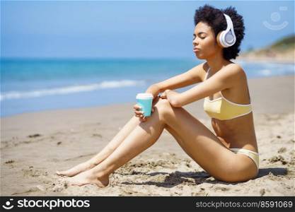 Black female sitting on the beach with wireless headphones, sipping a drink from a paper take-away cup.. African American girl sitting on the beach with headphones, sipping a drink from a take-away cup.
