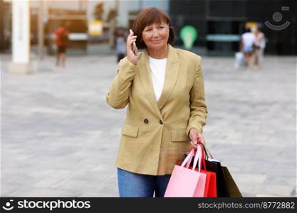 Black Friday. Smiling middle aged woman with bunch of shopping bags is communicating on cellphone by mall after shopping, concept of consumerism, sale, rich life. Commercial offer for advertising. Black Friday. Smiling middle aged woman with bunch of shopping bags is communicating on cellphone by mall after shopping, concept of consumerism, sale, rich life. Commercial offer for advertising.