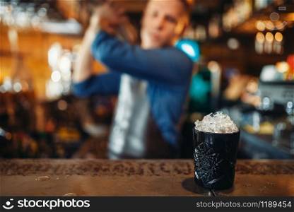 Black glass full of ice stands on bar counter, male barman with shaker on background. Barkeeper occupation, bartender job. Black glass full of ice stands on bar counter